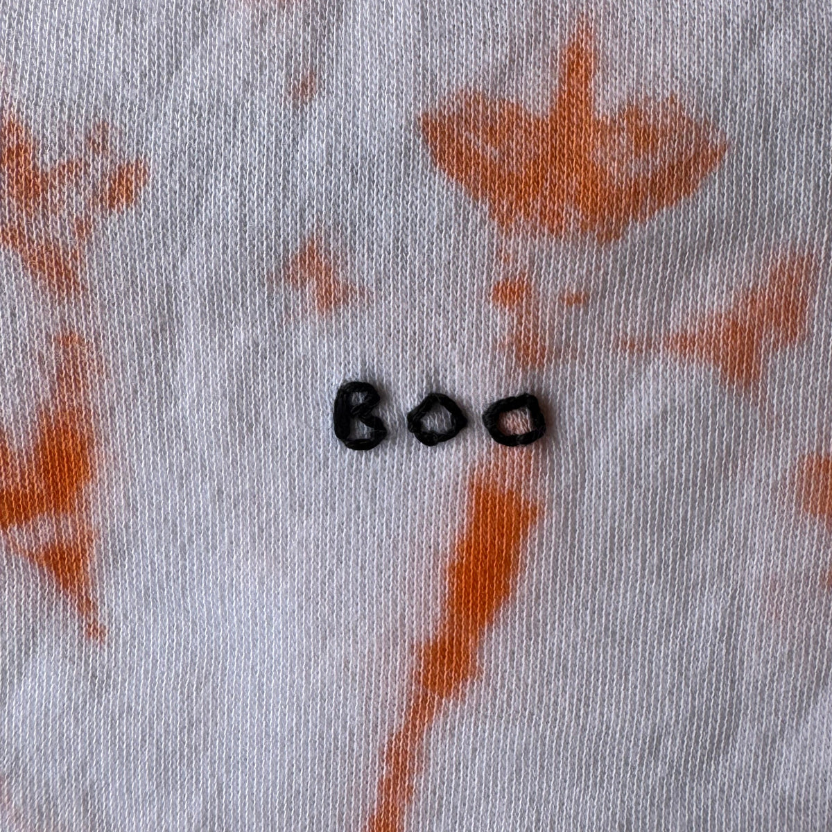 Close-up of an orange tie-dye onesie with hand embroidered BOO. 