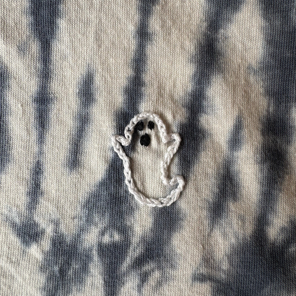 A small white embroidered ghost detail on a beige and midnight blue tie-dye cotton tee shirt.