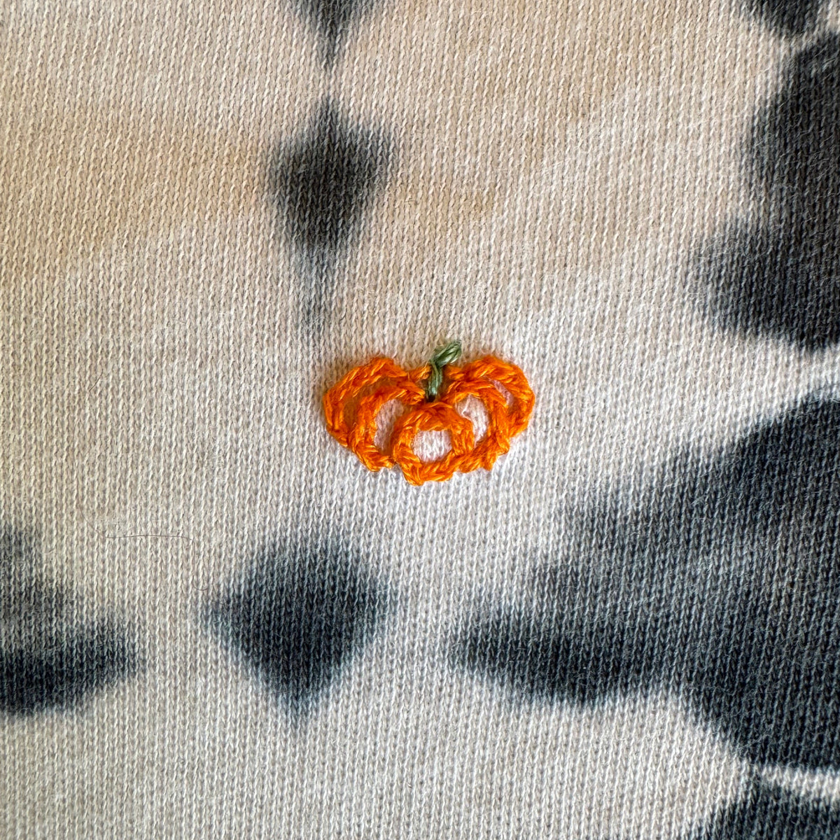 A close-up of a midnight blue and white cotton tie-dye infant onesie garment featuring a small orange hand embroidered pumpkin.