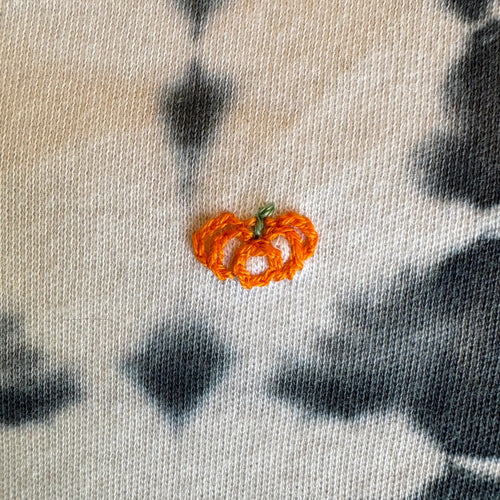 A close-up of a midnight blue and white cotton tie-dye infant onesie garment featuring a small orange hand embroidered pumpkin.