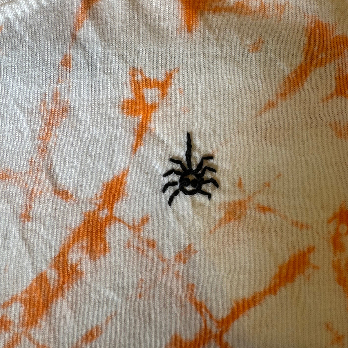 Close-up of a white t-shirt with orange tie-dye and a small black hand  embroidered spider on the chest.