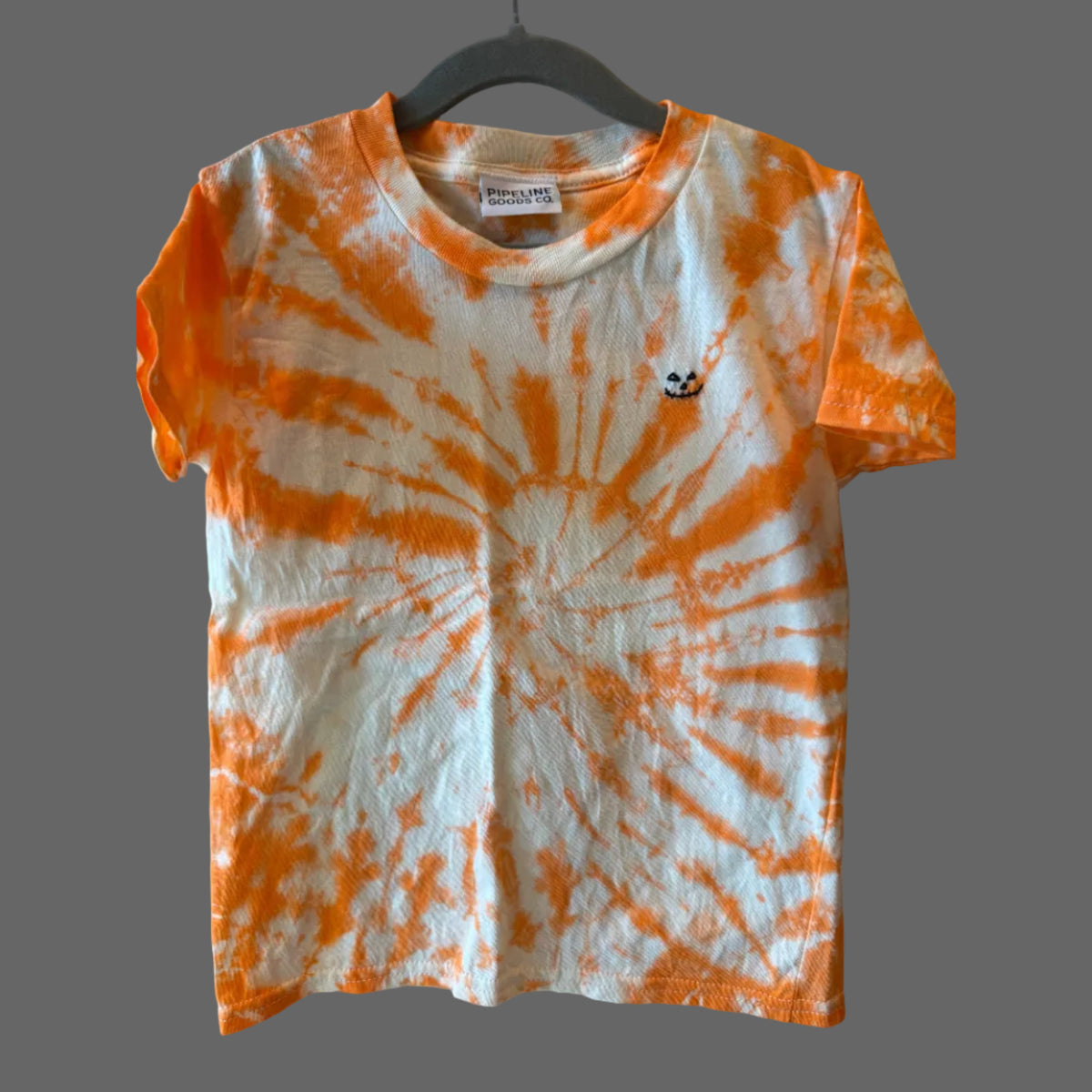 A short-sleeve cotton crewneck t-shirt with an orange and white tie-dye spiral pattern with a small black hand embroidered jack-o-lantern face on the left chest area.